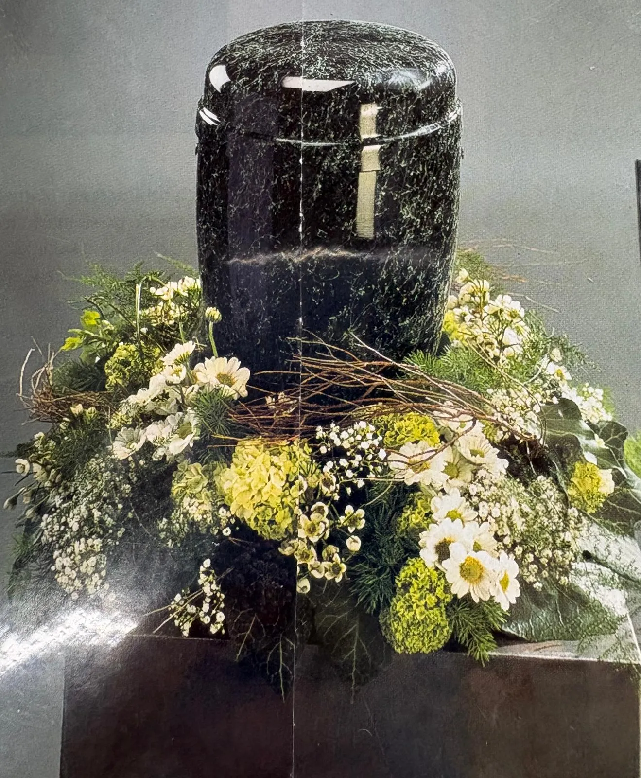 SC Corina Spriestersbach – Freie Rednerin – Urne aus schwarzem Marmor, umgeben von einem Blumenarrangement aus weißen Gänseblümchen und verschiedenen grünen und weißen Blättern.